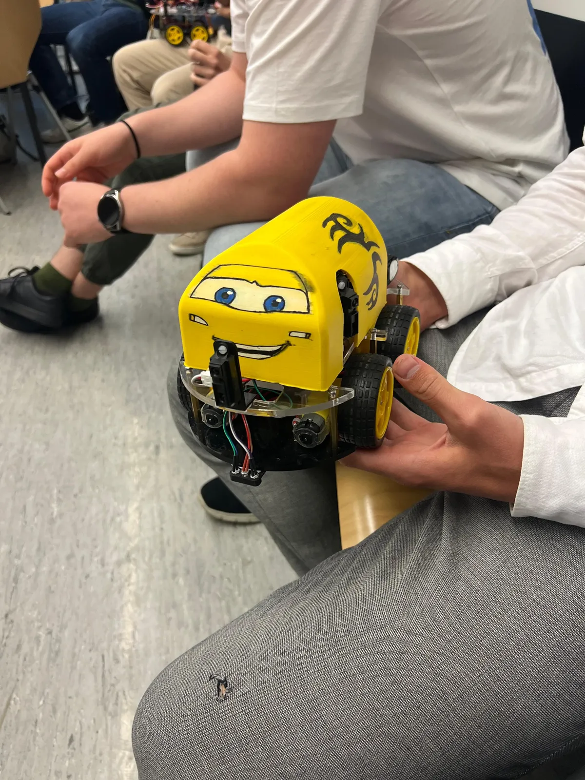 The robot held in two hands, with a yellow printed shell over the electronics and three distance sensors mounted at the front and sides.
