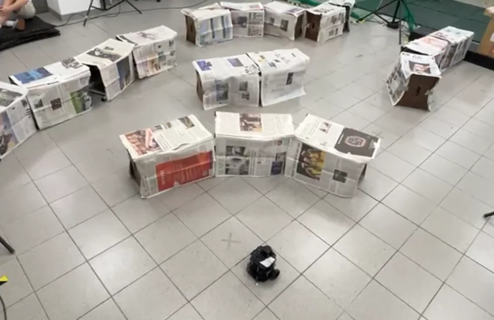 Photograph of a tiled lab floor with low cardboard boxes covered in newspaper arranged into a loose maze, and a small black TurtleBot sitting at a cross marked on the floor in the foreground.