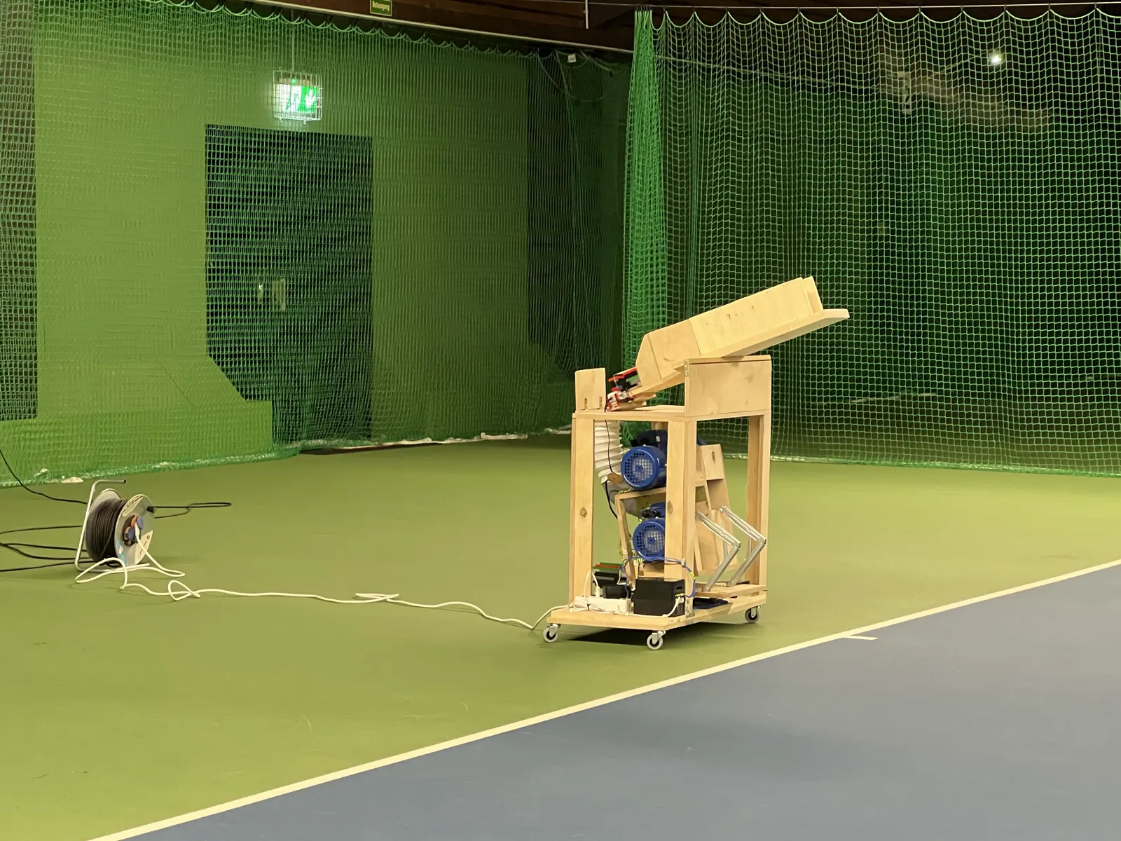 The finished tennis ball machine standing on an indoor court: a wooden frame on castors with a launcher chute angled upward, two blue motors inside, and a cable running off to the side.
